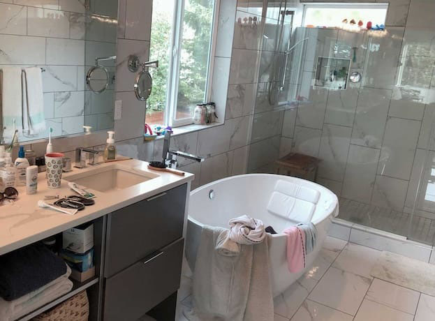 Modern master bathroom with marble-look tiles, glass shower doors, and a floating bathtub showing cluttered countertops and misplaced towels before professional cleaning in Bellevue.