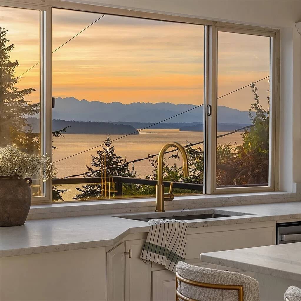 Clean kitchen sink with a brass faucet and marble counter below a window with a sunset view of Puget Sound