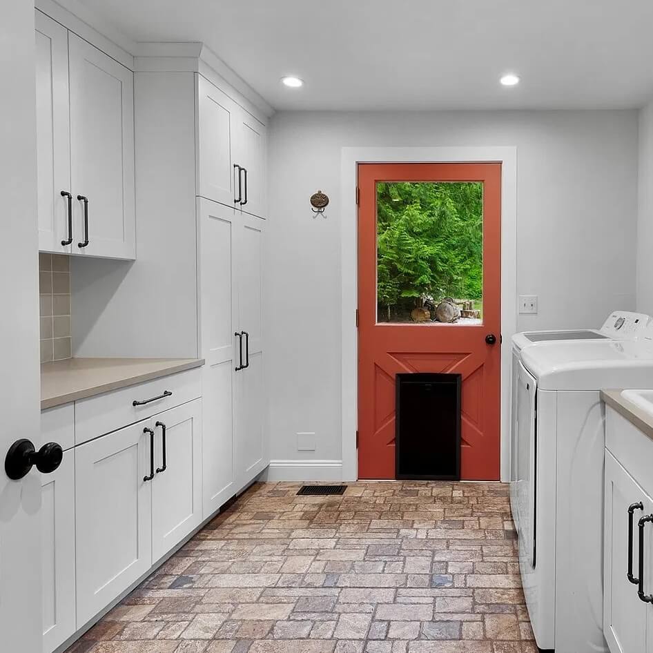 Clean mudroom and laundry room with white cabinets, brick floor and a red back door with a pet door