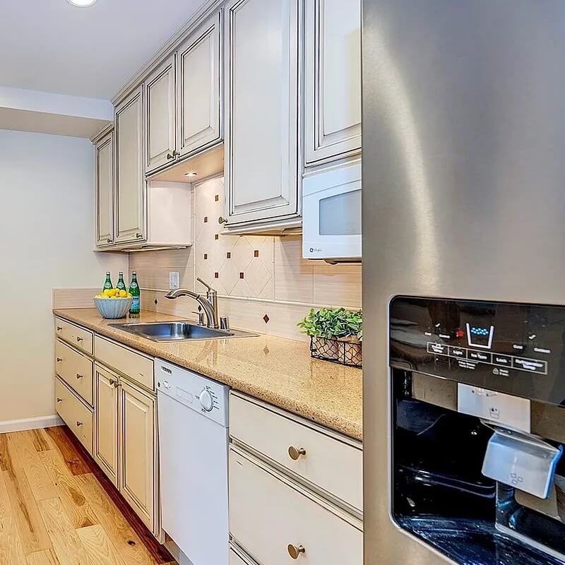 Freshly cleaned galley kitchen with cream cabinets, stone counters, stainless fridge and light hardwood floors
