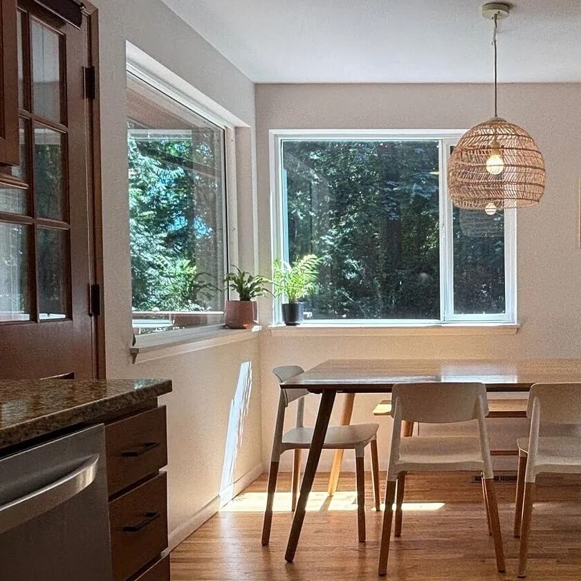 Sunny dining nook with a wood table, white chairs, hardwood floor and big windows looking out on trees
