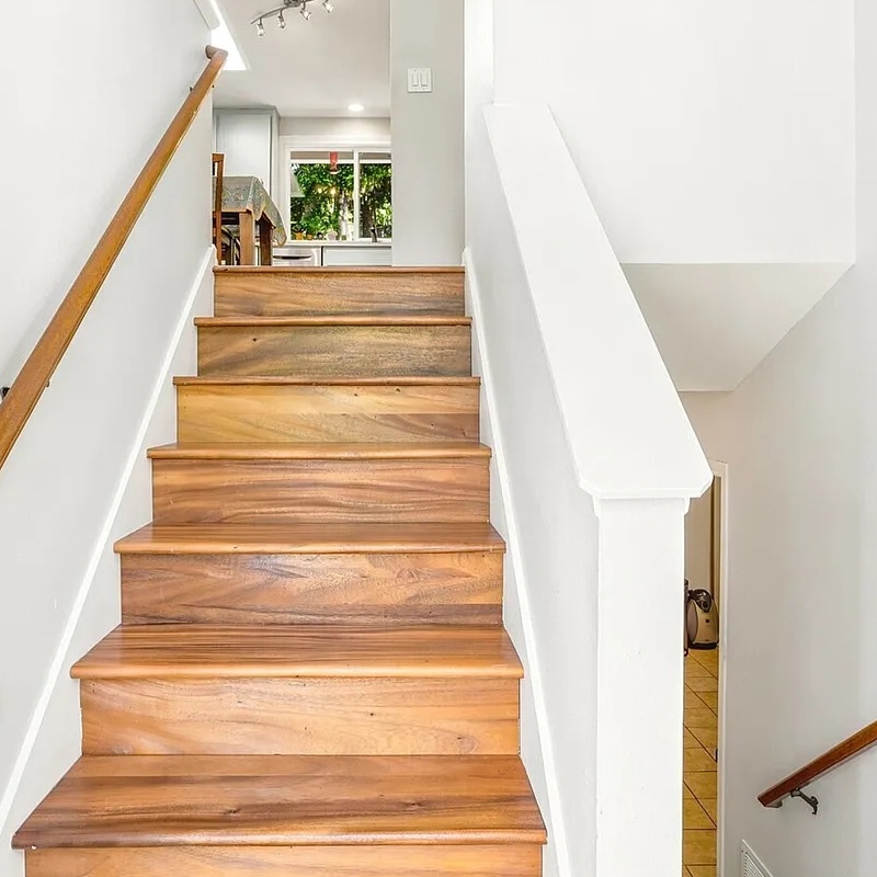 Straight hardwood staircase with white walls and a wood handrail leading up to a bright kitchen