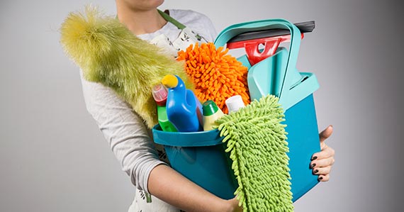 A slim white person with painted nails holding a bucket of cleaning supplies, including feather duster, squeegee, and various bottles of different colors.